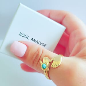 A gold plated Hug ring with turquoise stone worn on thumb also holding soul analyse ring box.