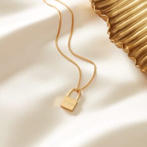 I AM LOVED gold padlock stainless steel necklace placed on a cream cloth with a gold plate alongside 