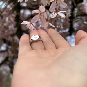 semi colon sterling silver oval signet ring worn on middle finger on a upright hand with blossom tree in background